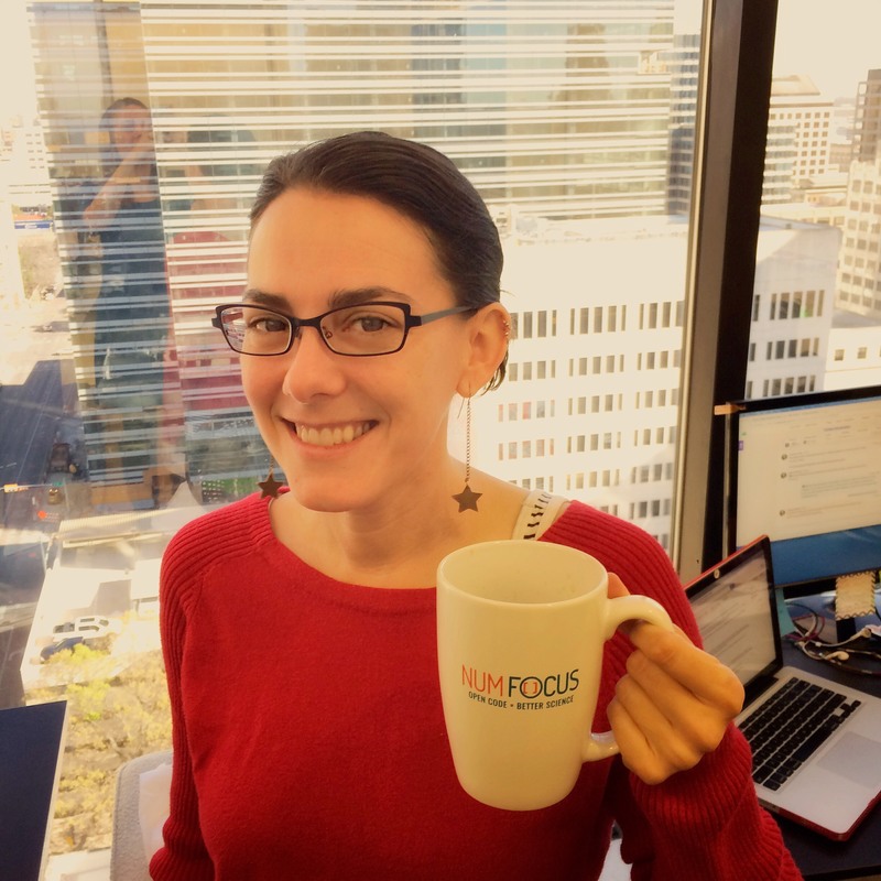 Gina Helfrich, Communications Director. Brunette woman with black wire frame glasses, smiling and holding a coffee cup that reads 